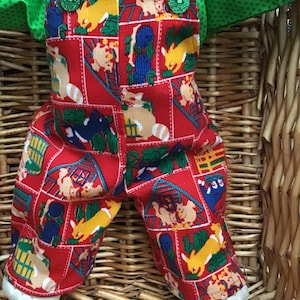 May include: Red overalls with a farm animal print, featuring a yellow, brown, and blue color scheme. The overalls have green buttons and are worn by a doll.