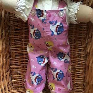 May include: A pink and blue baby shark print overall outfit for a doll. The overall has a white button closure and is worn over a white shirt with a lace trim.