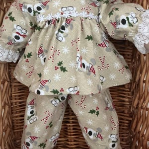 May include: A doll wearing a beige long-sleeved top and matching trousers featuring a repeating pattern of dogs in Santa hats, candy canes, holly, and white snowflakes. The top has a white lace collar and white lace trim on the sleeve cuffs.