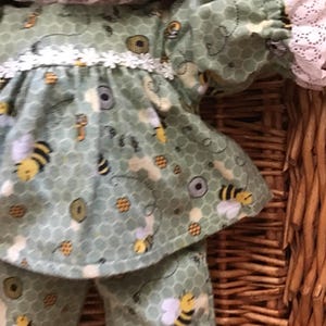 May include: A green and yellow doll outfit with a bee pattern. The outfit has a long-sleeved top with a white lace trim and matching trousers.