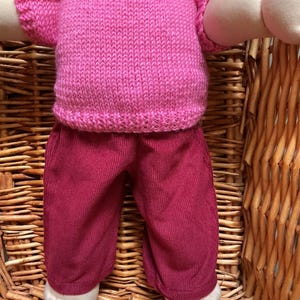 May include: A doll with brown hair wearing a short-sleeved, bright pink knitted shirt and dark maroon corduroy trousers. The doll has light-toned skin, visible arms, and bare feet, and is positioned in a seated posture with its legs and arms extended.