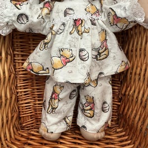 May include: A doll wearing a white long-sleeved top and matching trousers featuring a repeating Winnie the Pooh and honey pot print. The top includes a white lace collar, a white floral trim across the chest, and white lace cuffs on the sleeves.