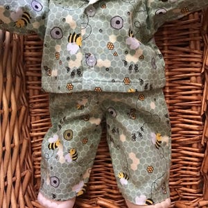 May include: A doll dressed in a light green bee-themed pajama set. The set features a honeycomb pattern and bee illustrations on the shirt and trousers. The doll is in a woven basket, and the outfit has a button-down shirt.