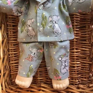 May include: A blue pyjama set with a koala print for a doll. The pyjamas have a button-up top and long trousers.
