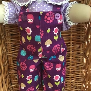May include: A doll dressed in a purple corduroy overall featuring a pattern of flowers, mushrooms, and owls. The overall has two purple buttons and is worn with a light purple polka dot shirt with a white lace collar and cuffs. The doll is in a wicker basket.