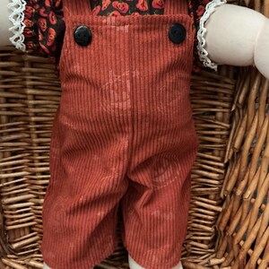 May include: A doll wearing rust-coloured corduroy overalls with a subtle pumpkin pattern and two black buttons at the chest. Underneath, the doll wears a black shirt featuring a repeating orange pumpkin print, a white lace collar, and lace cuffs.