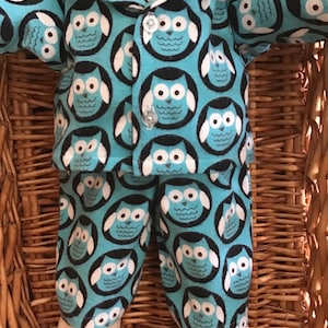 May include: A doll dressed in a blue owl-patterned pajama set. The set comprises a button-up shirt and trousers. The owl design has white and black details against a light blue backdrop. The doll's feet are visible, and the set is presented in a wicker basket.