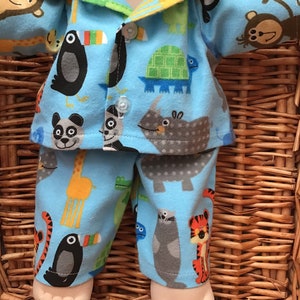 May include: A light blue pyjama set featuring a colourful animal print pattern including giraffes, toucans, turtles, pandas, rhinos, and tigers. The top is a button-down shirt with a collar, and the bottoms are matching trousers. The fabric has a soft texture.
