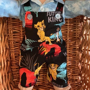 May include: A doll wearing a light blue collared shirt under black overalls featuring a colorful Lion King print. The fabric displays Simba, Timon, and Pumbaa silhouettes against a sunset background with the text Future King in white lettering.