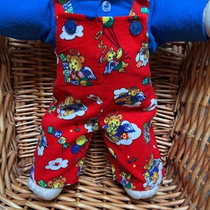 Peut inclure: Une poupée portant une chemise à col bleu vif sous une salopette en velours côtelé rouge. La salopette présente un motif répétitif d'ours en peluche ailés, de nuages et de ballons colorés. Le vêtement possède deux boutons bleu foncé.