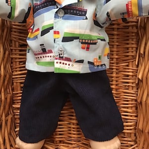 May include: A Cabbage Patch Kid doll wearing a blue and white button-down shirt with a ship pattern and dark blue corduroy trousers.