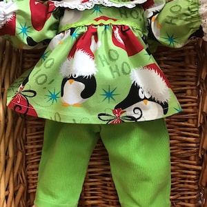 May include: A green and red Christmas-themed doll outfit with penguins and the words "Ho Ho" printed on it. The outfit includes a long-sleeved dress and matching pants.