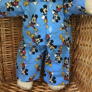 May include: A doll wearing a light blue long-sleeved button-down shirt and matching trousers featuring an all-over print of Mickey Mouse in various poses. The shirt has a collar and two clear buttons visible on the front placket of the garment.