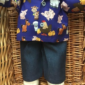 May include: A doll wearing a blue button-down shirt patterned with farm animals, including pigs, cows, and sheep. The doll is also wearing dark blue denim pants and white shoes. The doll is sitting in a woven basket.
