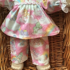 May include: A doll dressed in a pink and white Care Bears outfit. The outfit comprises a top with a frilled collar and sleeves, and matching trousers. The fabric displays Care Bears, rainbows, and clouds.