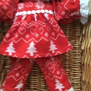 May include: A red and white Christmas-themed doll outfit with a white lace trim. The outfit features a long-sleeved top and pants with a repeating pattern of hearts, snowflakes, and Christmas trees.
