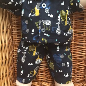 May include: A dark blue pyjama set with a yellow and blue construction vehicle print. The pyjamas have a button-up shirt and long trousers.