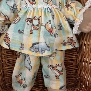 May include: A doll wearing a long-sleeved top and trousers set featuring a light blue and yellow plaid pattern with illustrations of Winnie the Pooh, Tigger, and Eeyore. The top has a white lace collar and white lace trim on the sleeve cuffs.