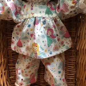 May include: A white doll wearing a pink and white fairy-themed pyjama set with a white lace trim. The doll is sitting in a wicker basket.