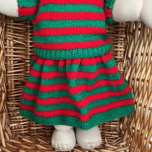 May include: A doll with dark brown hair wearing a hand-knitted short-sleeved dress featuring horizontal red and green stripes. The doll has a light-toned fabric body with visible toes and is positioned within a woven wicker basket structure.