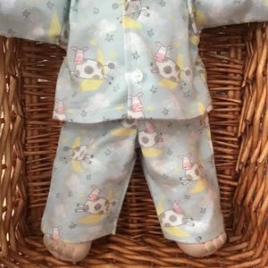 May include: Pale blue pyjama set with a cow and moon design. The set comprises a button-up shirt and matching trousers. The fabric displays a repeating pattern of cows, moons, and stars.