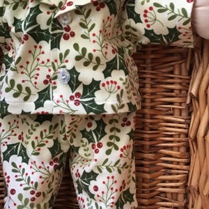 May include: A doll wearing a cream-colored pajama set with a green holly and red berry pattern. The pajamas have a button-up collar and long sleeves.