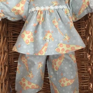 May include: A doll wearing a light blue pajama set with a giraffe pattern. The pajamas include a top with a white lace collar and a matching pair of pants. The giraffe pattern features yellow and orange spots. The doll is in a wicker basket.