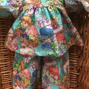 Peut inclure: Poupée vintage vêtue d'une tenue bleu clair à imprimé Fraise. La tenue comprend une robe à volants et des manches, ainsi qu'un pantalon assorti. L'imprimé présente des personnages de Fraise, des fraises et des fleurs.