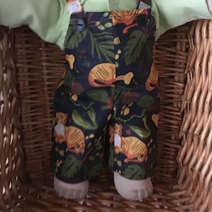 May include: A doll dressed in a light green shirt and dark blue dungarees. The dungarees have a tiger and leaf pattern in orange, yellow, and green. The doll is in a wicker basket.