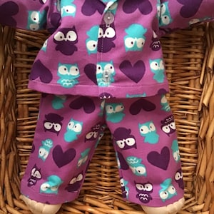 May include: Purple pajamas with a pattern of owls and hearts for a doll. The pajamas have a button-up top and long pants.