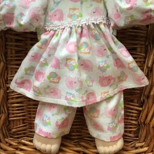 May include: A doll wearing a white and pink patterned outfit featuring elephants and owls. The outfit includes a dress with a ruffled hem and sleeves, and matching pants. The doll is in a woven basket.