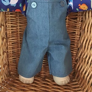 May include: A doll wearing blue denim overalls with two light blue glittery buttons at the top, layered over a dark blue collared shirt featuring a colorful pattern of various fish in shades of orange, white, and light blue with green accents.