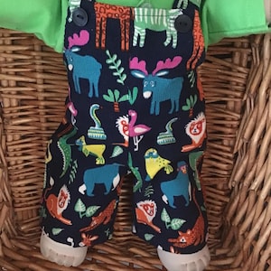 May include: A pair of dark blue corduroy dungarees featuring a colourful animal print pattern including moose, lions, snakes, and flamingos. The dungarees have two black buttons at the top of the straps and are worn over a bright lime green collared shirt.