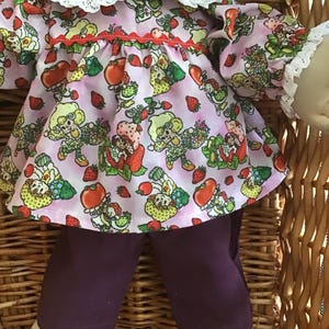 May include: A doll wearing a pink dress with a strawberry and character print, white lace trim, and purple leggings. The doll is in a woven basket. The dress has a red trim around the neckline.