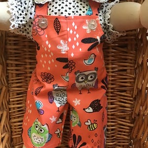 May include: A doll dressed in a coral jumpsuit featuring an owl and floral design. The jumpsuit has decorative clear buttons and is worn with a white top with a black heart pattern and lace trim. The doll is in a wicker basket.
