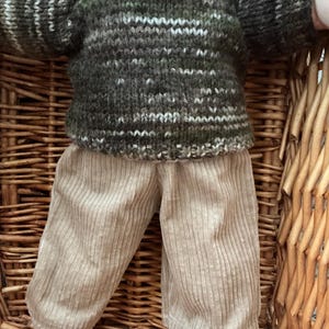 May include: A doll wearing a long-sleeved, crew-neck jumper featuring a variegated knit pattern in shades of dark green, olive, and white, paired with light beige corduroy trousers that have a vertical ribbed texture and a straight-leg cut.