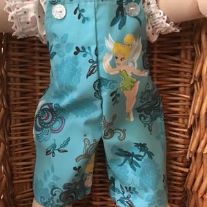 May include: Doll's clothing set. The light blue dungarees feature a floral and dragonfly design, and a Tinkerbell graphic. The dungarees have silver buttons and are paired with a white dotted blouse with ruffled sleeves.