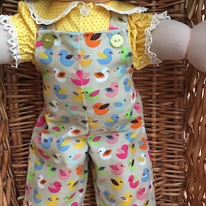 May include: A doll wearing a yellow short-sleeved shirt with a white polka dot pattern and white lace trim on the collar and sleeves, paired with beige corduroy overalls featuring a colourful repeating print of small ducks in blue, pink, orange, and green.