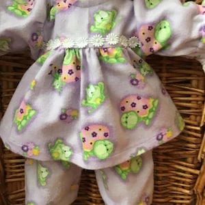 Puede incluir: Muñeca vestida con un conjunto de forro polar lavanda con un estampado de tortugas. El conjunto incluye una parte superior de manga larga con cuello de encaje blanco y una falda, además de pantalones a juego. El diseño de la tortuga presenta tortugas verdes con detalles rosas y morados.