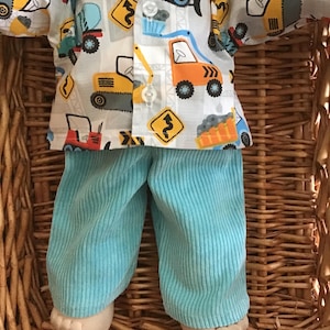 May include: A doll dressed in light blue corduroy trousers and a button-up shirt patterned with construction vehicles. The shirt displays diggers, trucks, and road signs on a light grey backdrop. The doll is in a wicker basket.