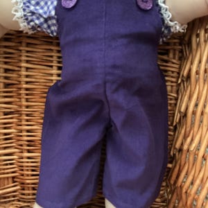 May include: A doll wearing a purple pair of dungarees with two purple buttons at the chest, layered over a blue and white gingham shirt featuring short sleeves with white lace trim and a white collar with a single button at the centre.
