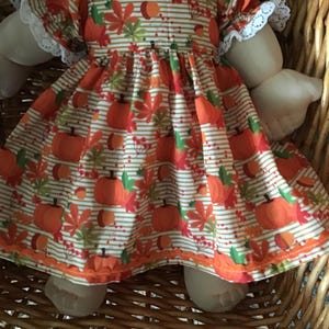 May include: A white dress with orange and brown pumpkins, leaves, and stripes. The dress has a lace trim on the sleeves.