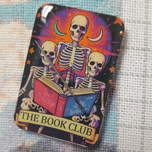 May include: A rectangular needle minder featuring a glossy, glittery illustration of three skeletons reading open books. The design includes a purple and orange background with crescent moons and stars. The text THE BOOK CLUB is printed at the bottom.