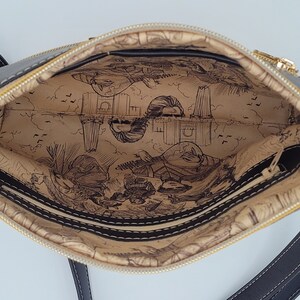 May include: Black crossbody bag with a gold zipper and a beige lining featuring a printed design of a city skyline and people.