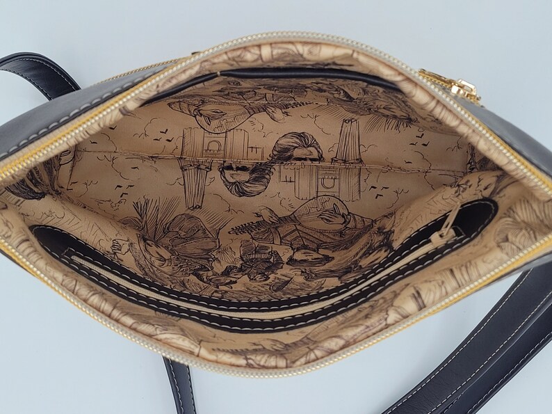May include: Black crossbody bag with a gold zipper and a beige lining featuring a printed design of a city skyline and people.