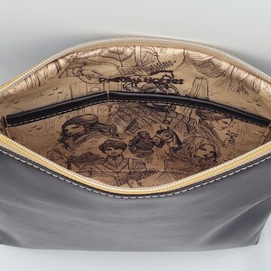 May include: A black leather zippered pouch with a gold zipper and a cream lining. The lining features a printed design of a fantasy scene with characters and a horse.