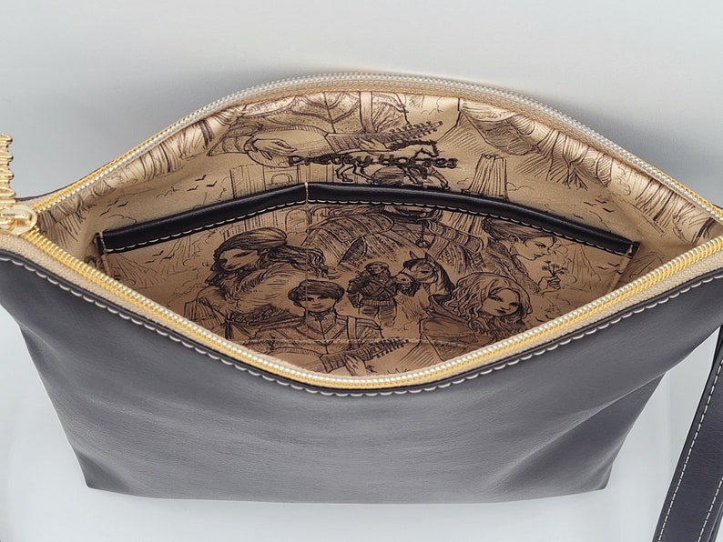May include: A black leather zippered pouch with a gold zipper and a cream lining. The lining features a printed design of a fantasy scene with characters and a horse.