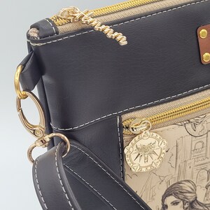 May include: A black faux leather crossbody bag with a gold zipper and a gold charm featuring a wolf's head. The bag has a beige canvas lining with a printed design of a woman and a city.