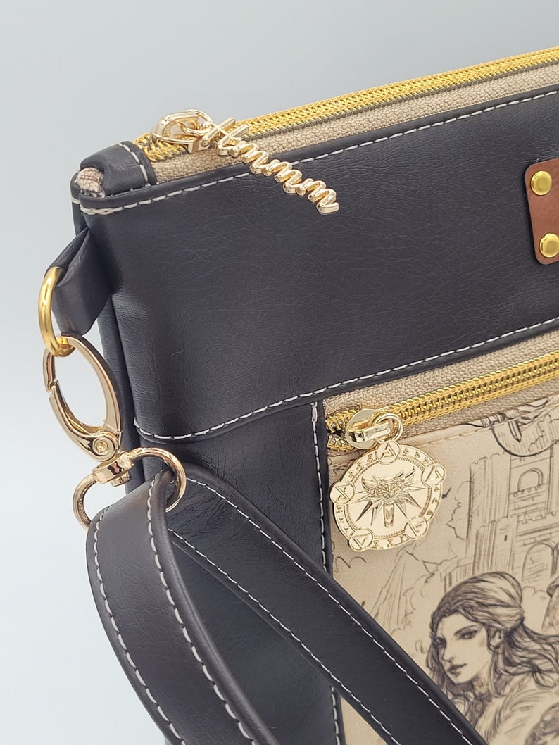 May include: A black faux leather crossbody bag with a gold zipper and a gold charm featuring a wolf's head. The bag has a beige canvas lining with a printed design of a woman and a city.