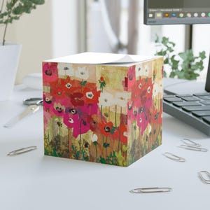 May include: A square memo cube featuring a vibrant floral print of red, pink, and white poppies on a textured, multi-colored background. The paper block has a flat top and vertical sides, displaying a repeating artistic pattern of blooming flowers.